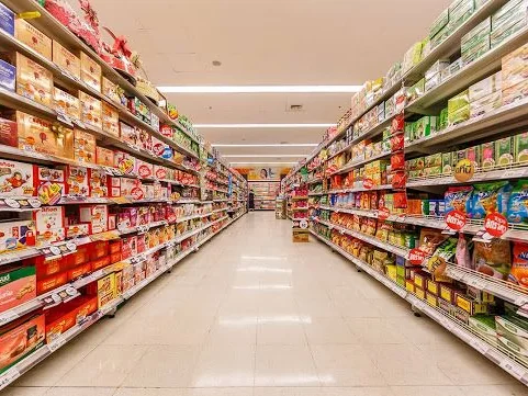 Long, brightly lit supermarket aisle with fully stocked shelves of packaged foods on both sides